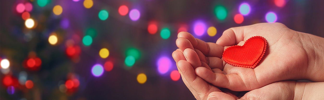 Festive background with a person’s cupped hands with a red heart representing giving during the holidays.